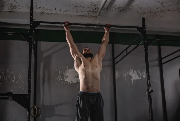 Mann hängt entspannt an einer Klimmzugstange in einem Fitnessraum. Darstellung der Übung zur Förderung der Schultergesundheit, Verbesserung der Mobilität und Stärkung der Rotatorenmanschette