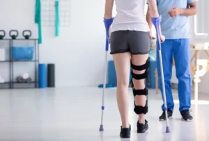 Eine Patientin mit Knieorthese und Unterarmgehstützen wird in einer modernen Physiotherapie-Praxis bei der Krankengymnastik nach Operationen begleitet. Der Therapeut unterstützt sie bei den ersten Mobilisationsschritten, um Beweglichkeit und Stabilität nach der OP wiederherzustellen. Im Hintergrund sind verschiedene Trainingsgeräte für die Rehabilitation zu sehen.