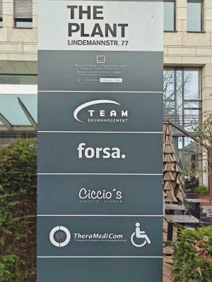 Außenstele "The Plant" an der Lindemannstraße 77. Wegweiser zu Physiotherapie TheraMediCom mit Rollstuhl-Symbol für den barrierefreien Zugang.